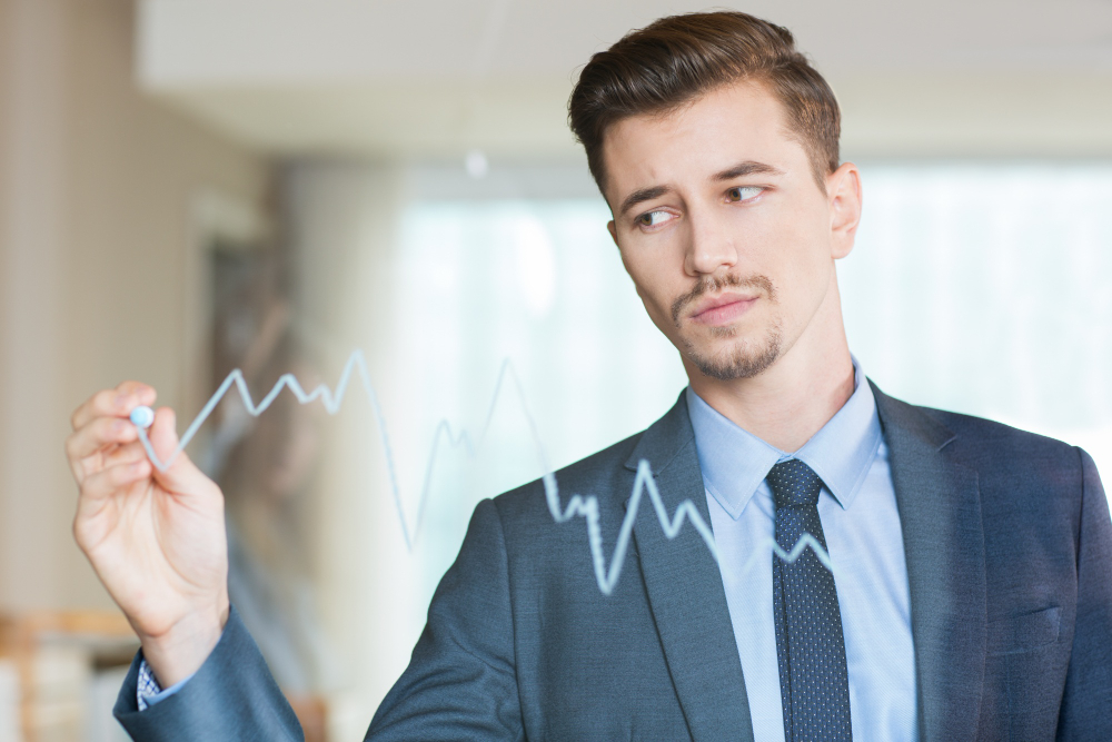 Businessman in a suit drawing a fluctuating financial line graph on transparent glass, illustrating investment analysis.
