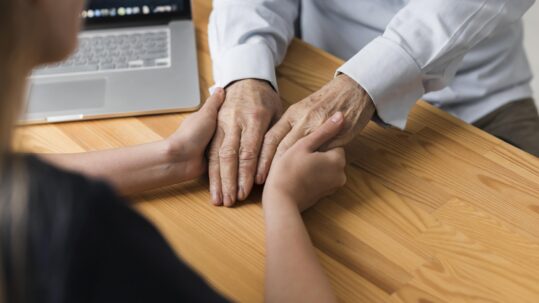 Person gently holding an older adult's hands across a table, illustrating support and planning for long-term care needs.