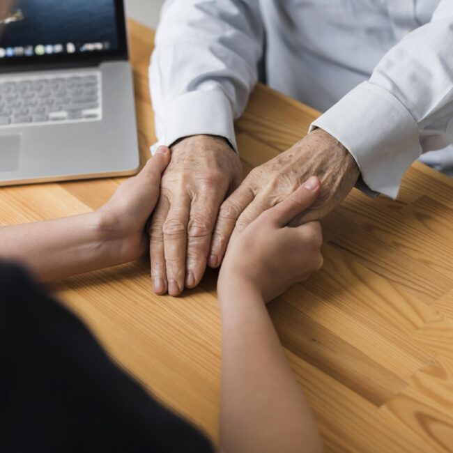 Person gently holding an older adult's hands across a table, illustrating support and planning for long-term care needs.