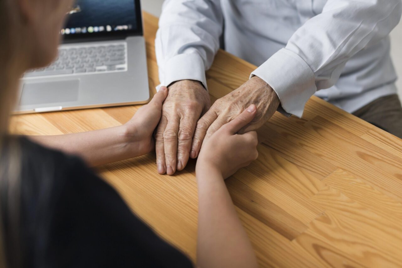 Person gently holding an older adult's hands across a table, illustrating support and planning for long-term care needs.