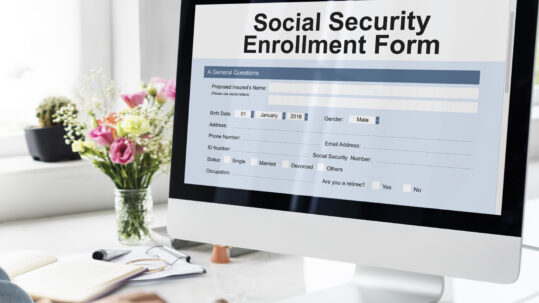 Person using a computer displaying a Social Security enrollment form while reviewing retirement benefit information.