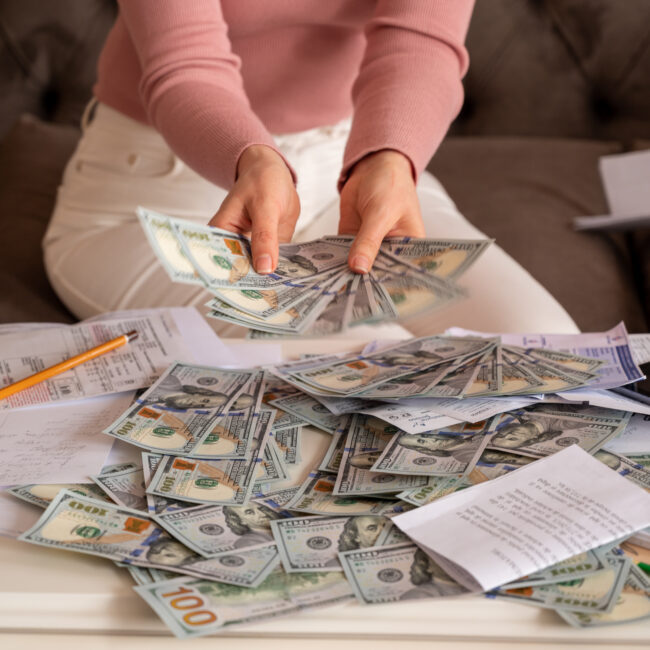 Woman counting U.S. dollar bills at a table covered with cash, receipts, and financial paperwork.