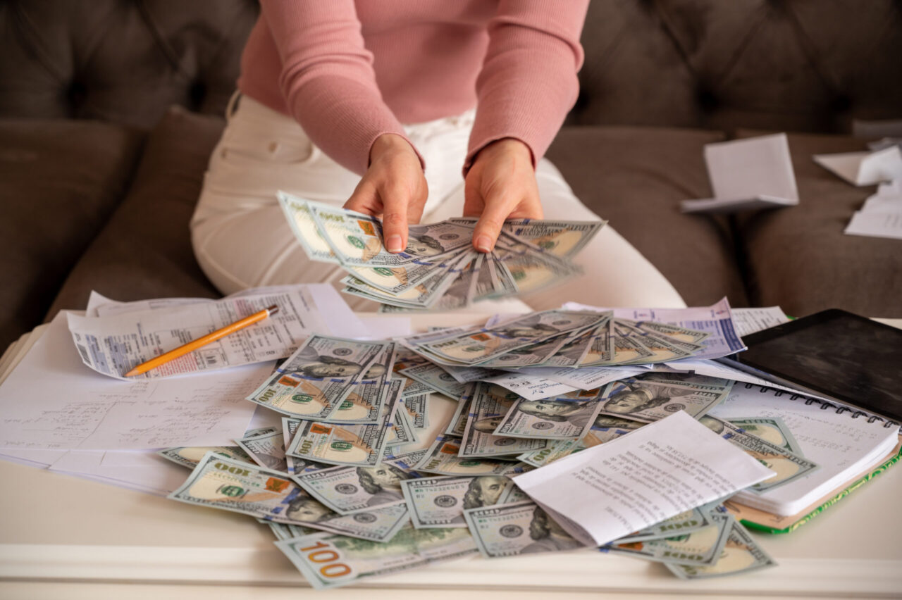 Woman counting U.S. dollar bills at a table covered with cash, receipts, and financial paperwork.