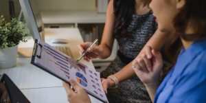 Two Asian business women collaborating in an office, reviewing a financial report with charts and graphs, pointing at data